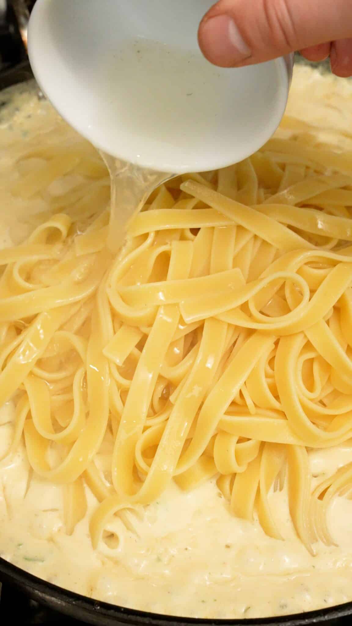 Fettuccine noodles and pasta water being added to alfredo sauce.