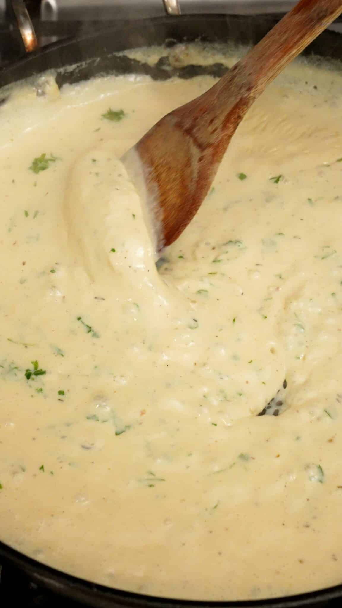 A wooden spatula stirring fresh parsley into alfredo sauce.