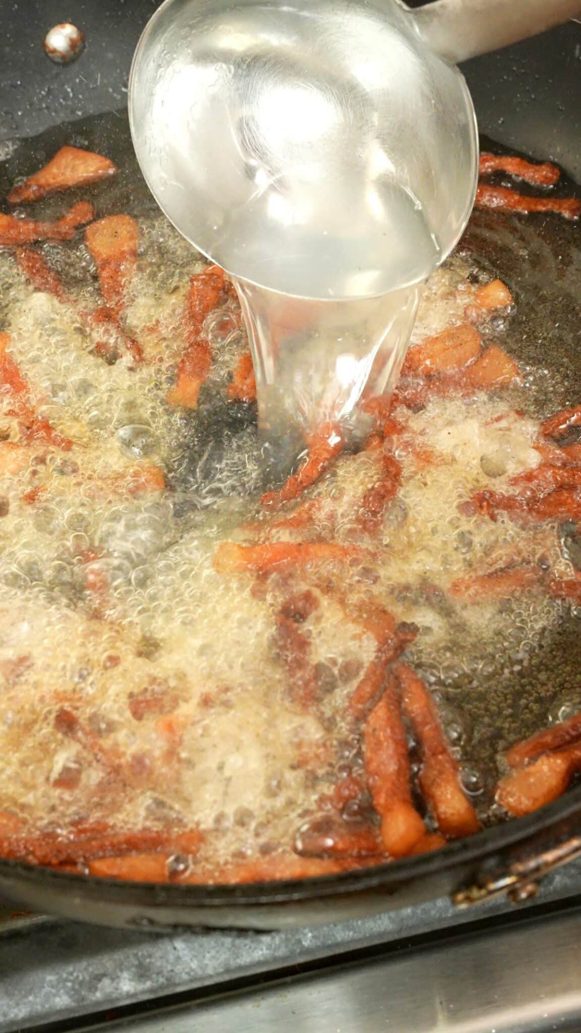 Pasta water being ladled into a pan with guanciale and fat.