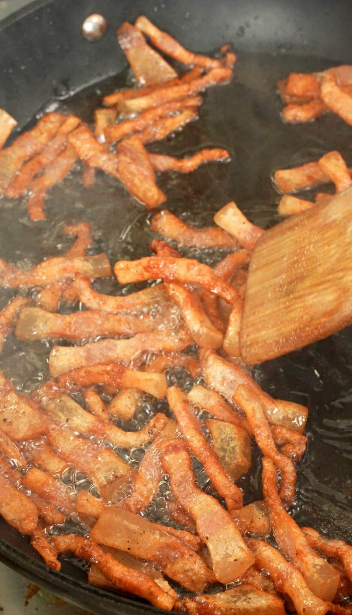 A wooden spatula mixing guanciale in a pan.
