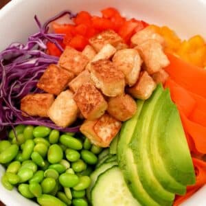 Buddha bowl in a bowl with bell pepper, carrot, avocado, edamame, crispy tofu, and red cabbage.
