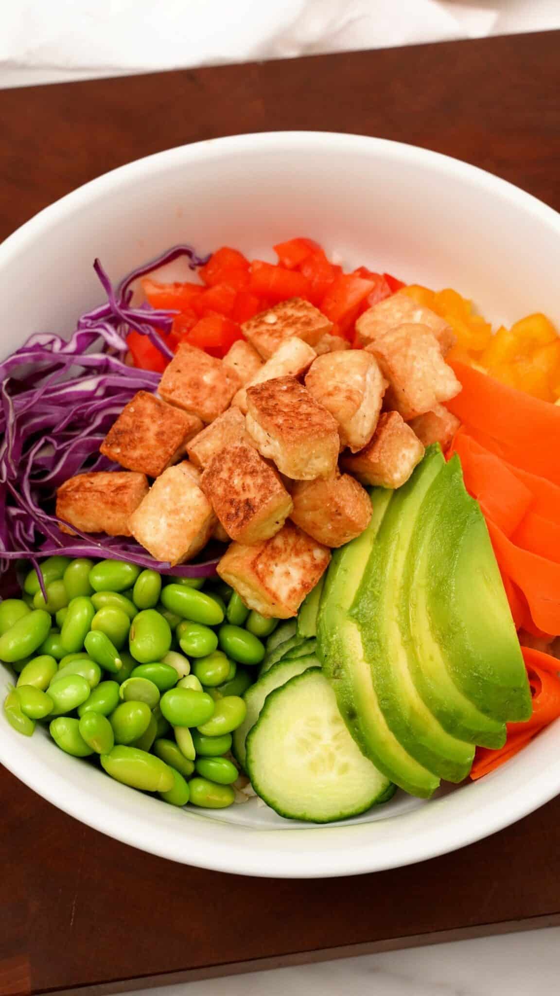 Buddha bowl in a bowl with bell pepper, carrot, avocado, edamame, crispy tofu, and red cabbage.
