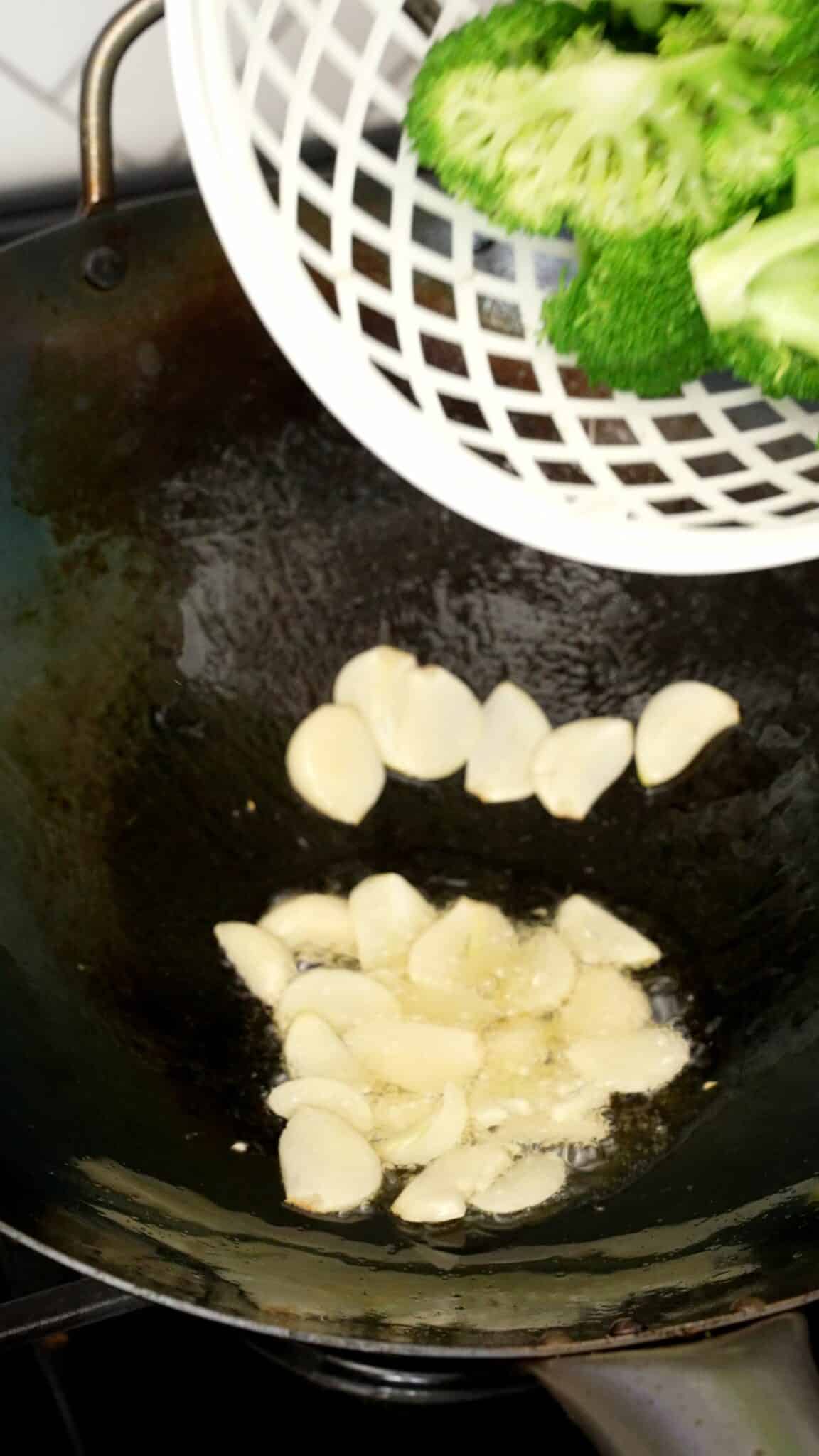 Sliced garlic cooking in oil in a wok with broccoli being added to the wok.
