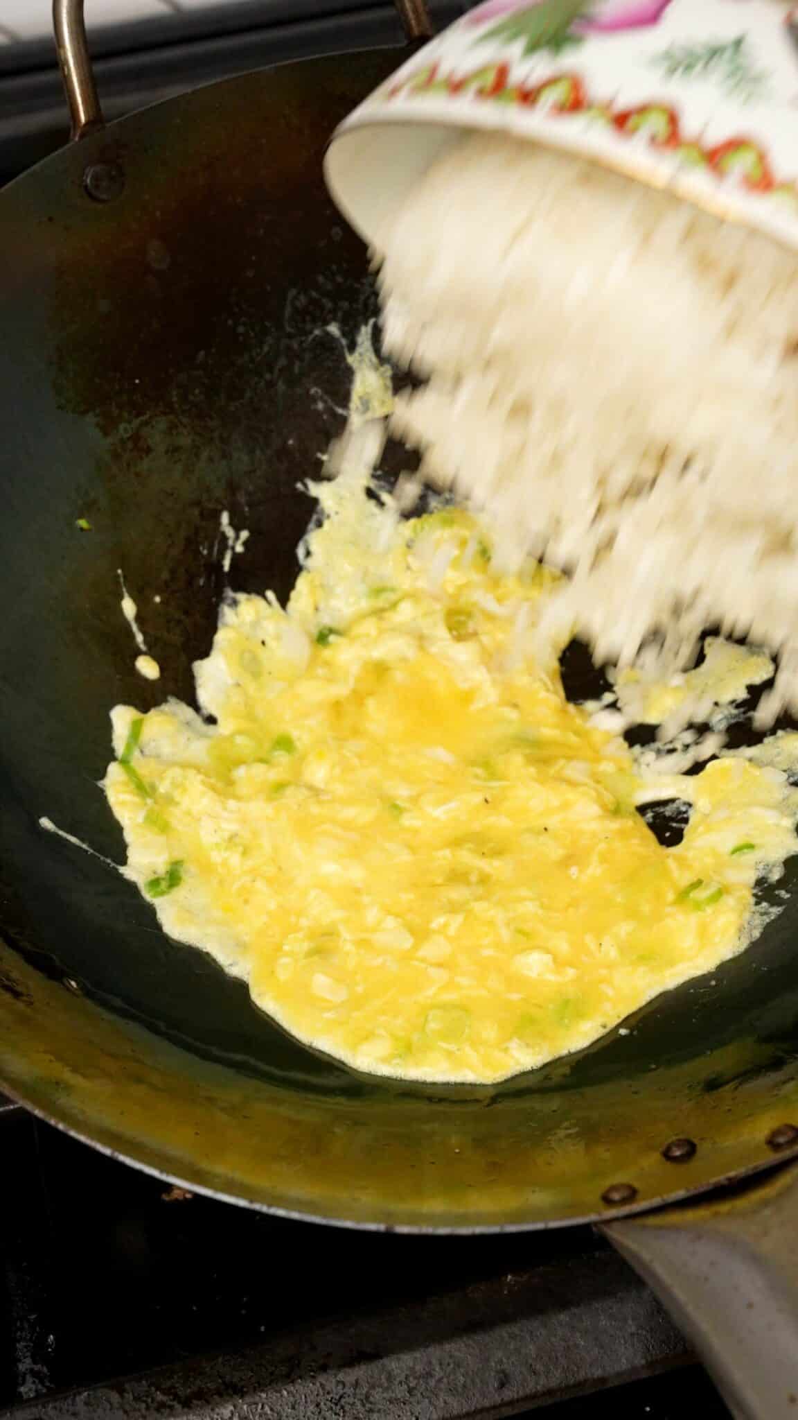 Slightly cooked beaten egg with scallion whites in a wok with white rice being added in.