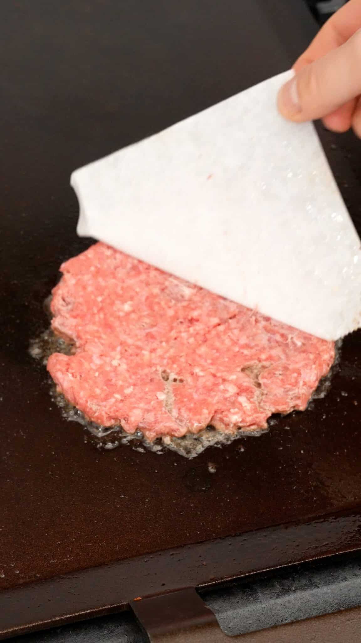 A hand removing the parchment paper from the smashed burger patty.