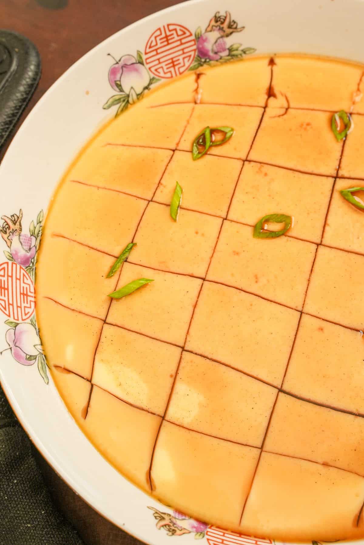 Chinese steamed eggs in a bowl with scallions and soy sauce cut into squares.