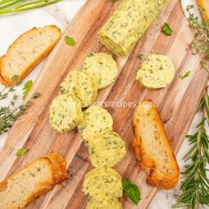 Garlic herb compound butter on a cutting board with herbs and bread. The butter is cut into circles.