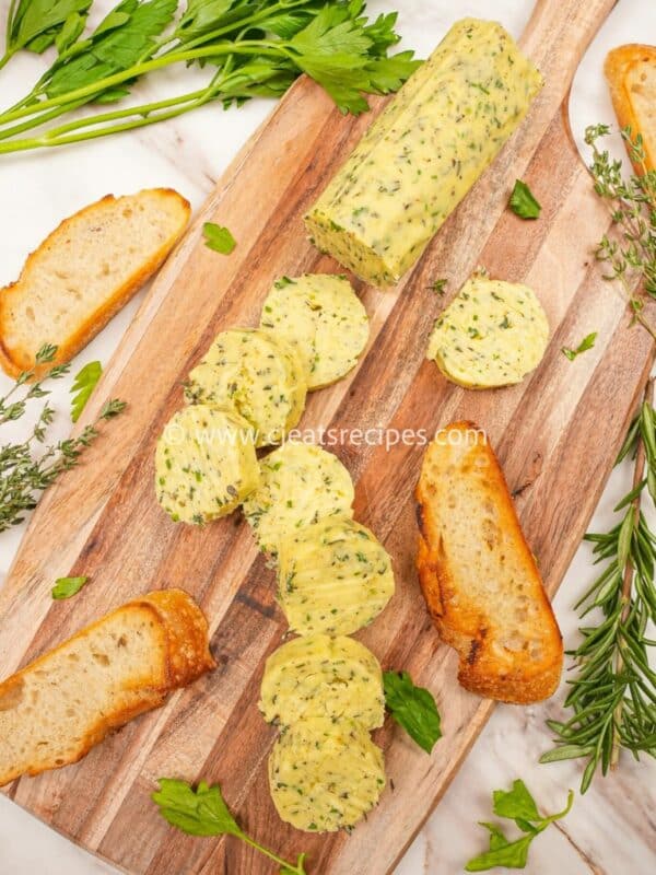 Garlic herb compound butter on a cutting board with herbs and bread. The butter is cut into circles.