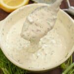 A bowl of tartar sauce with a spoon next to lemon and fresh dill.