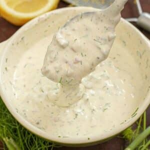 A bowl of tartar sauce with a spoon next to lemon and fresh dill.