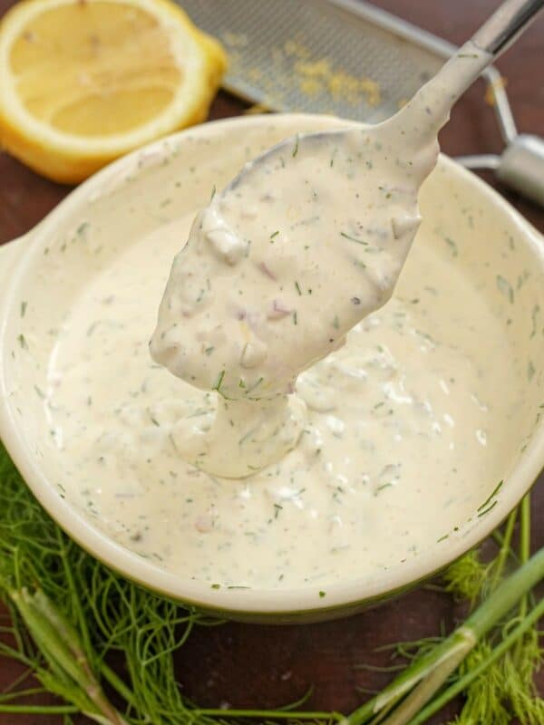 A bowl of tartar sauce with a spoon next to lemon and fresh dill.