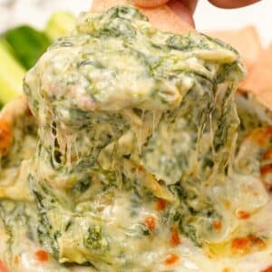 A hand scooping spinach artichoke dip with a tortilla chip.
