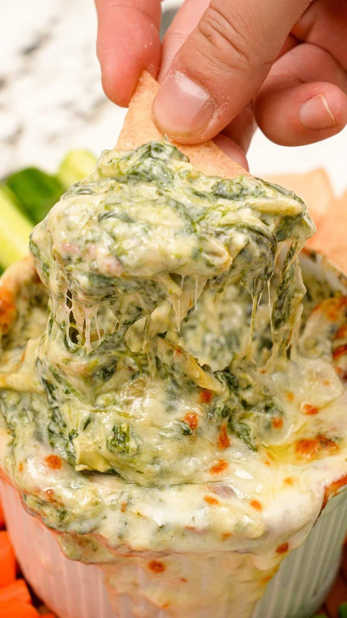A hand scooping spinach artichoke dip with a tortilla chip.