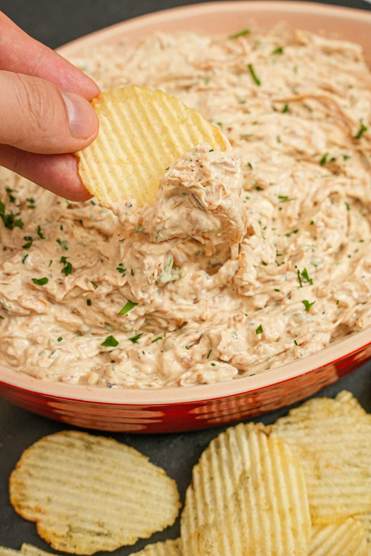 A hand scooping Caramelized Onion Dip with thick cut potato chips.