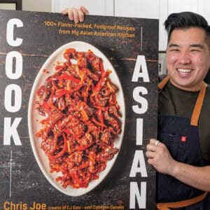 Chris Joe, author of COOK ASIAN, holding up a blow up cover of his debut cookbook in the kitchen.