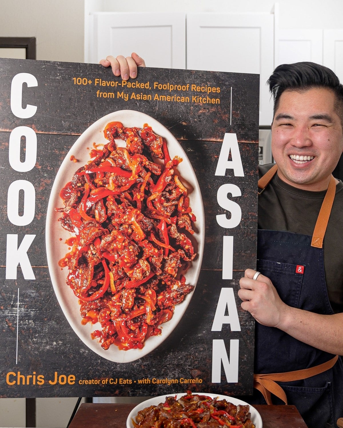 Chris Joe, author of COOK ASIAN, holding up a blow up cover of his debut cookbook in the kitchen.