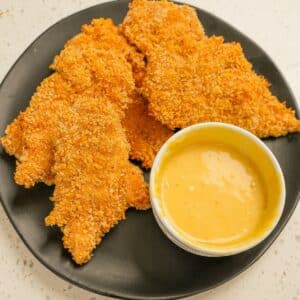 A plate of crispy baked chicken tenders on a black plate with honey mustard sauce.