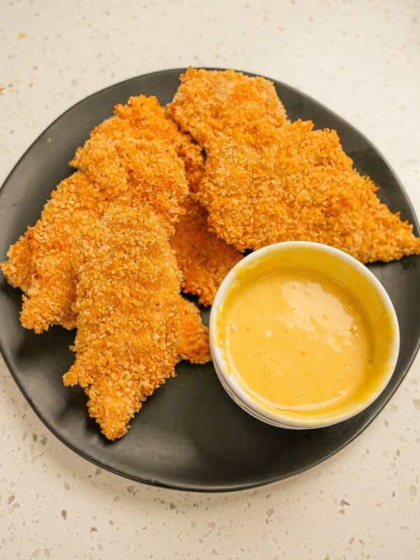 A plate of crispy baked chicken tenders on a black plate with honey mustard sauce.