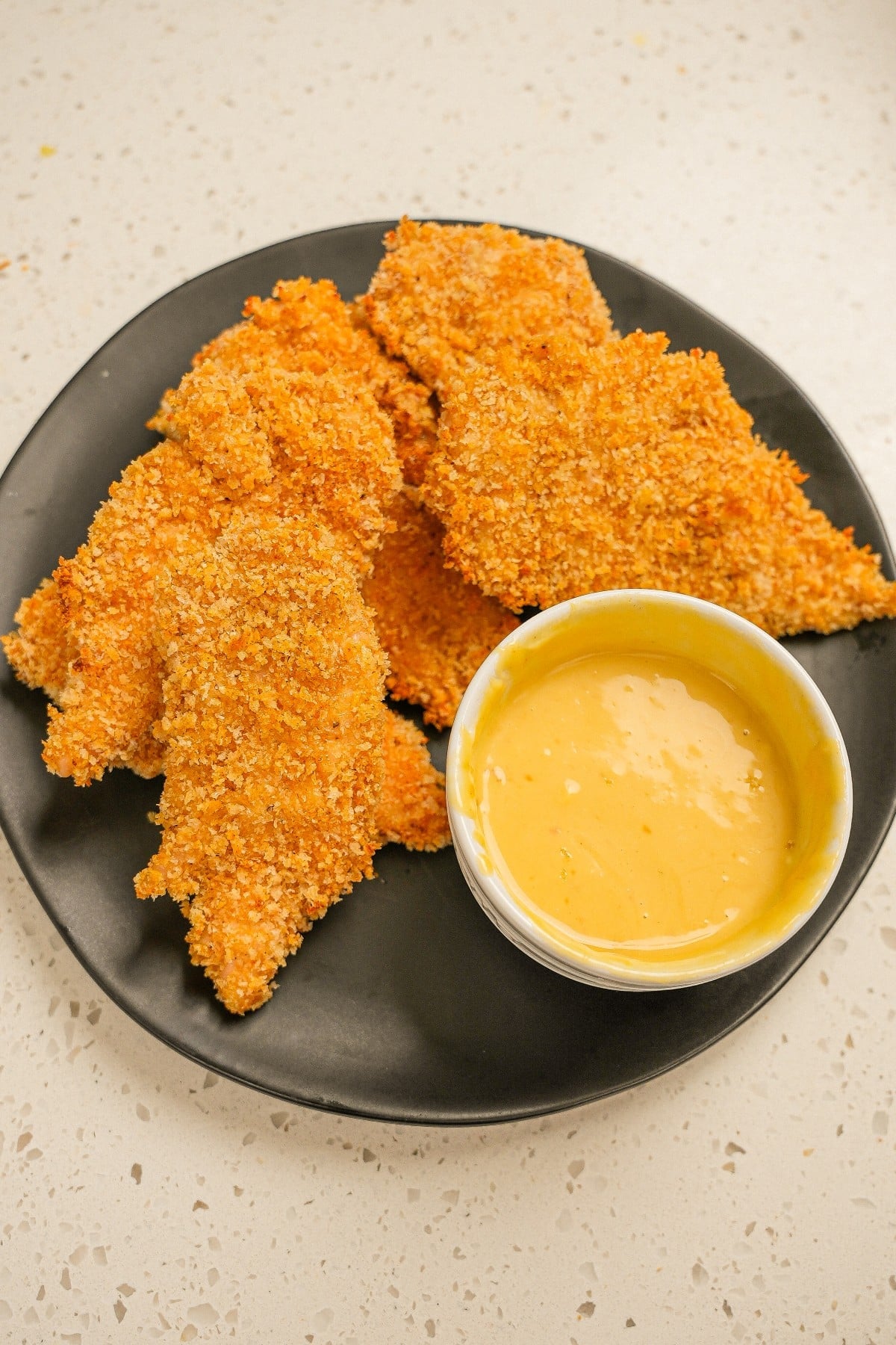 A plate of crispy baked chicken tenders on a black plate with honey mustard sauce.
